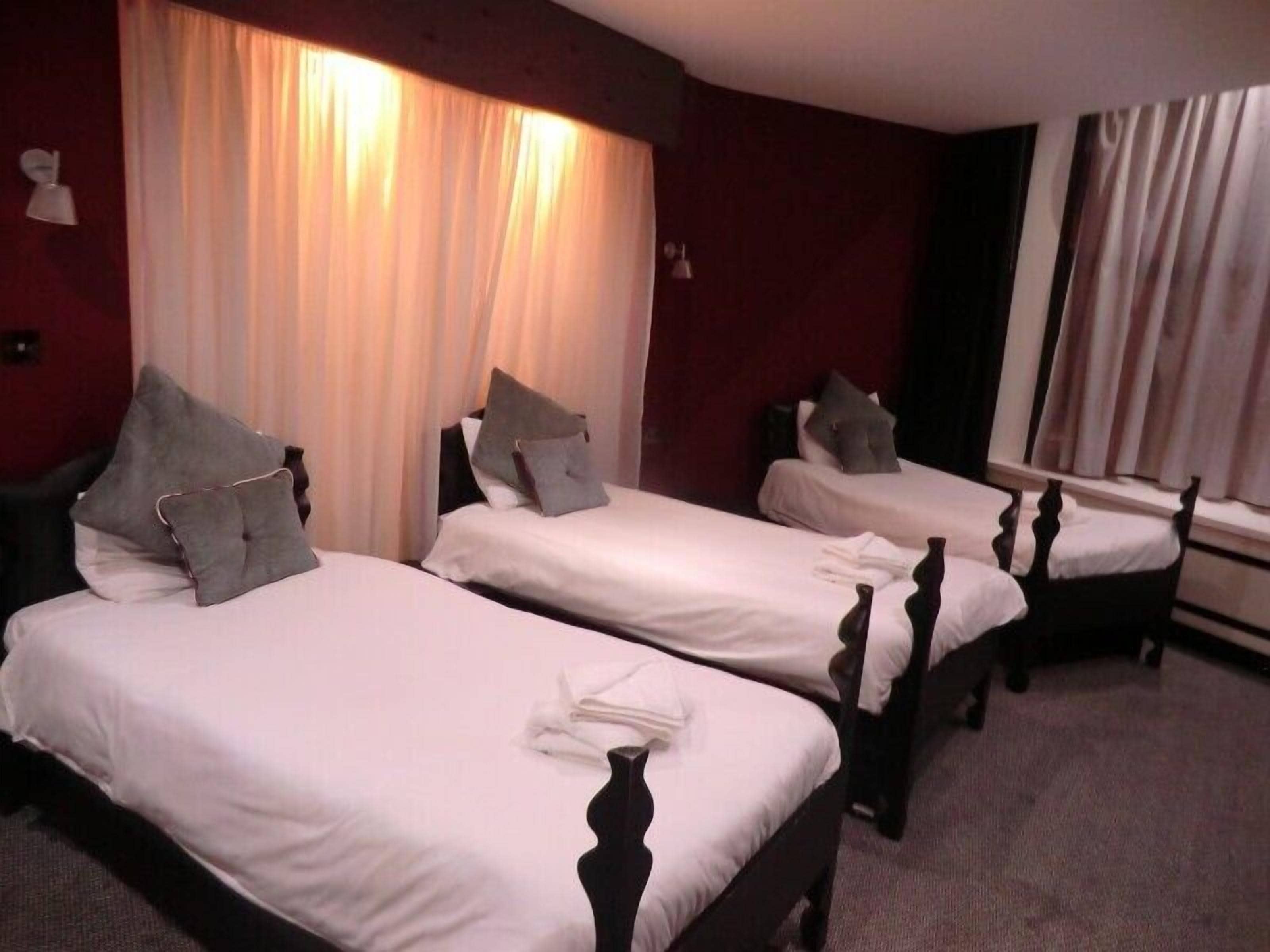 A hotel room with three single beds, each with a wooden headboard and white bedding. There are curtains on the windows, and a small table with a lamp and a chair.