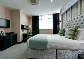 Habitación de hotel moderna con una cama grande, un televisor de pantalla plana, un escritorio con lámpara y una ventana con cortinas.