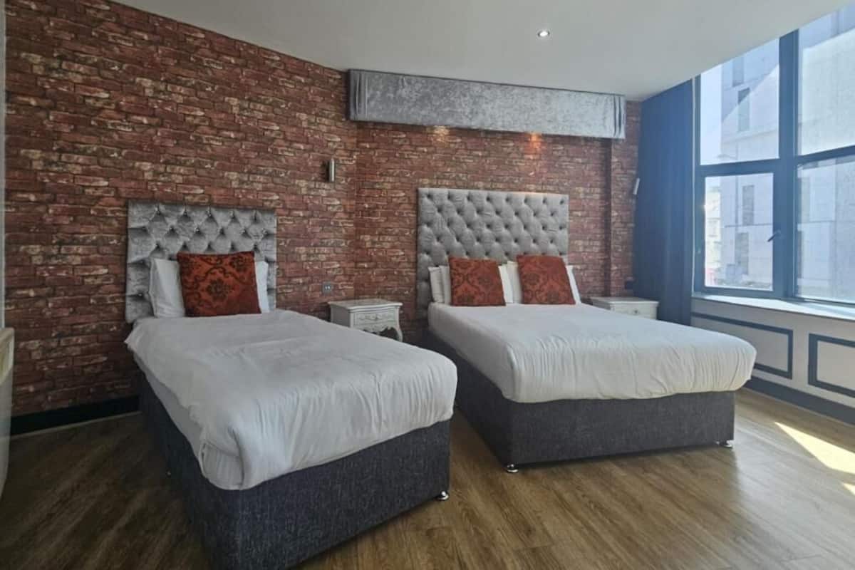 Hotellihuone, jossa on kaksi sänkyä, tiiliseinä, suuri ikkuna ja moderni kattorakenne.