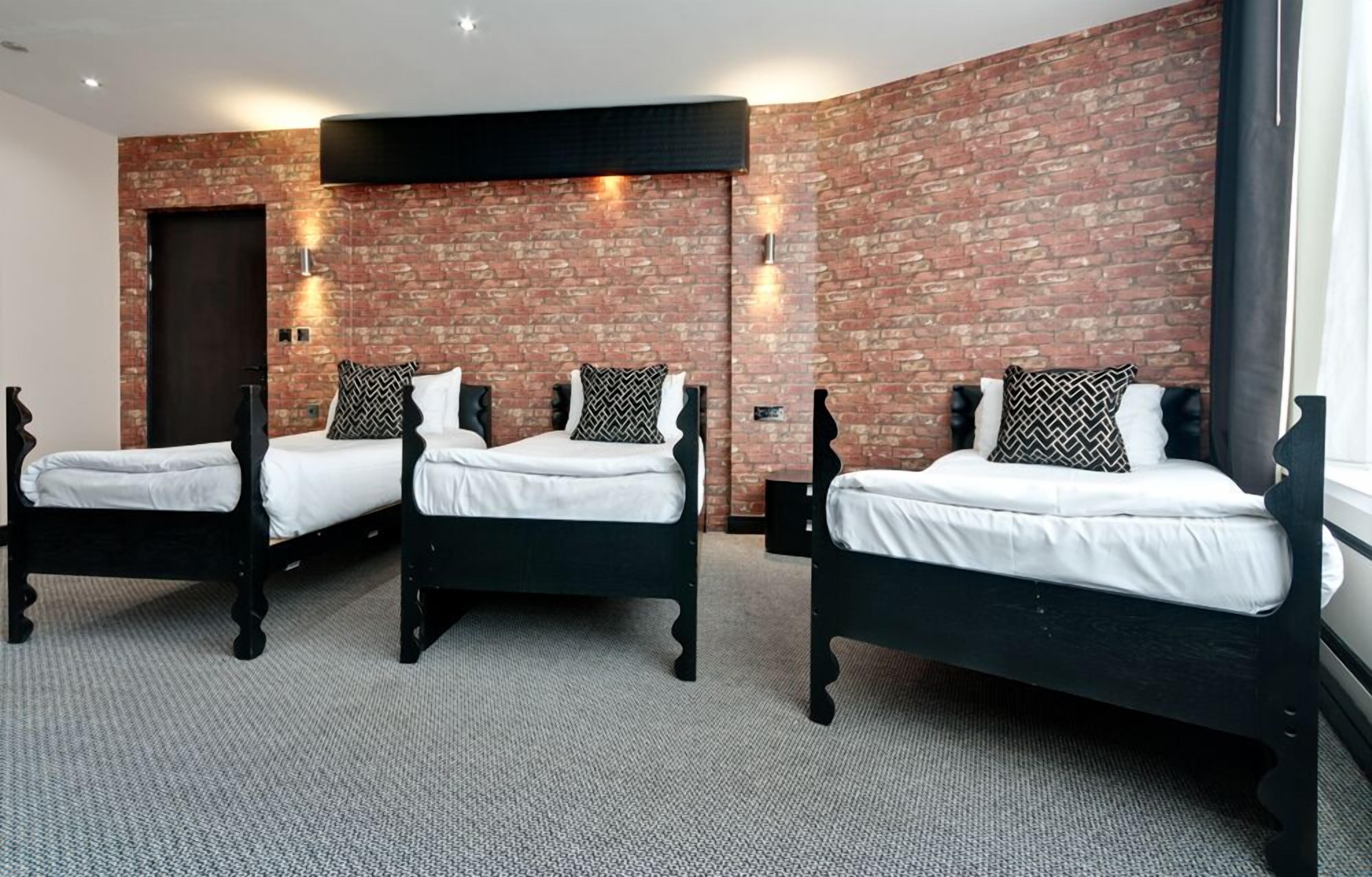 A hotel room with three black beds, white bedding, and a brick wall.