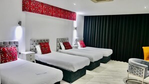 A hotel room with three beds, each with a grey headboard and white bedding. There is a red patterned curtain on the window, a grey chair, and a white nightstand with a lamp. - Printworks Hotel (Liverpool)