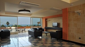 A spacious indoor area with a checkered floor, a seating area with sofas and a table, a television mounted on a brick wall, and large windows offering a view of the sea.
