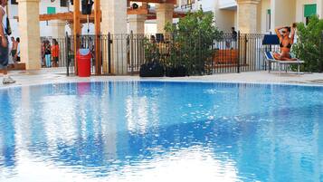 A swimming pool with a wooden pergola, a red trash bin, and a fenced area with people.