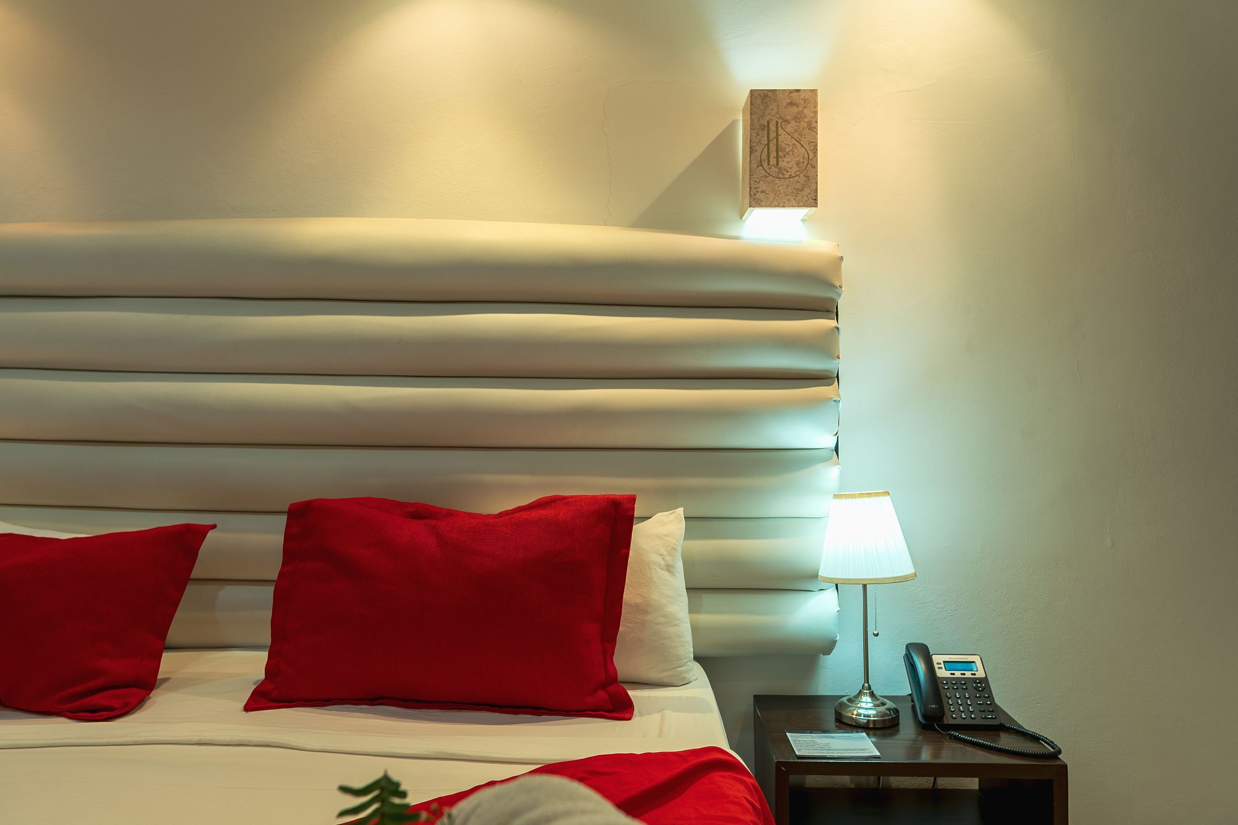 A hotel room with a neatly made bed, red and white pillows, a bedside lamp, and a telephone on a nightstand.