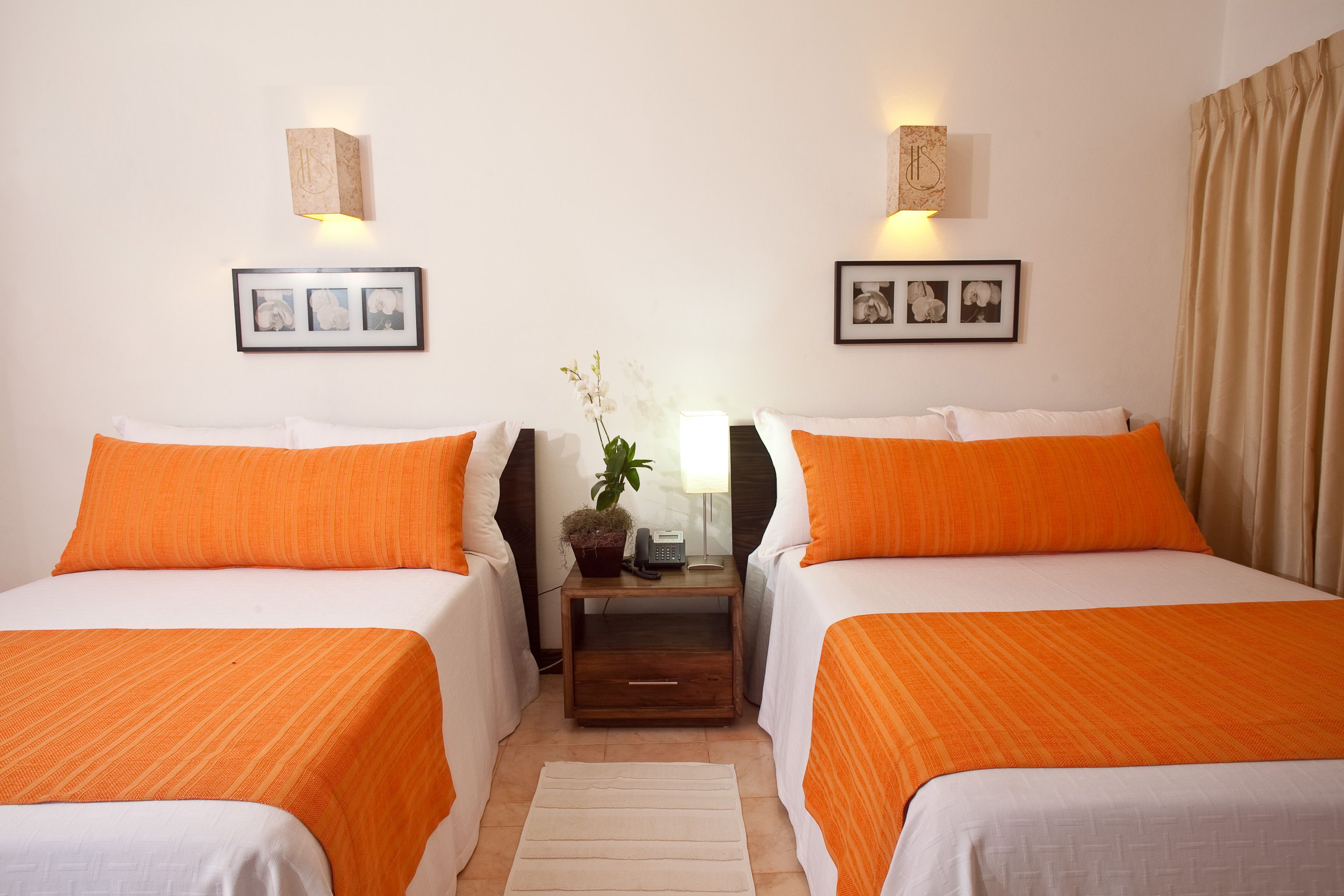 A hotel room with two beds, each with an orange bedspread, a nightstand with a lamp and a plant, and framed pictures on the walls.