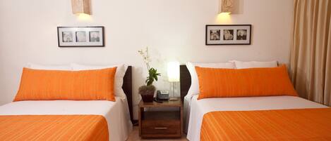 A hotel room with two beds, each with an orange bedspread, a nightstand with a lamp and a plant, and framed pictures on the walls.