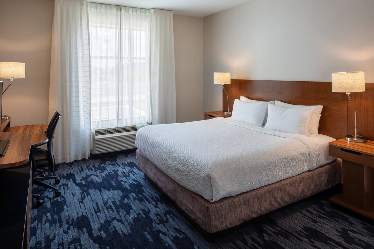 A hotel room with a large bed, two bedside lamps, a desk with a chair, and a window with curtains.