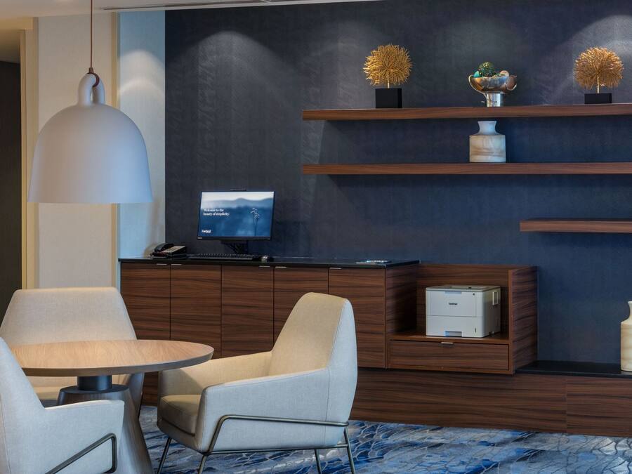 Una habitación de hotel moderna con una pared oscura, estantes de madera, una mesa redonda, dos sillas, un escritorio con una impresora y un televisor montado.