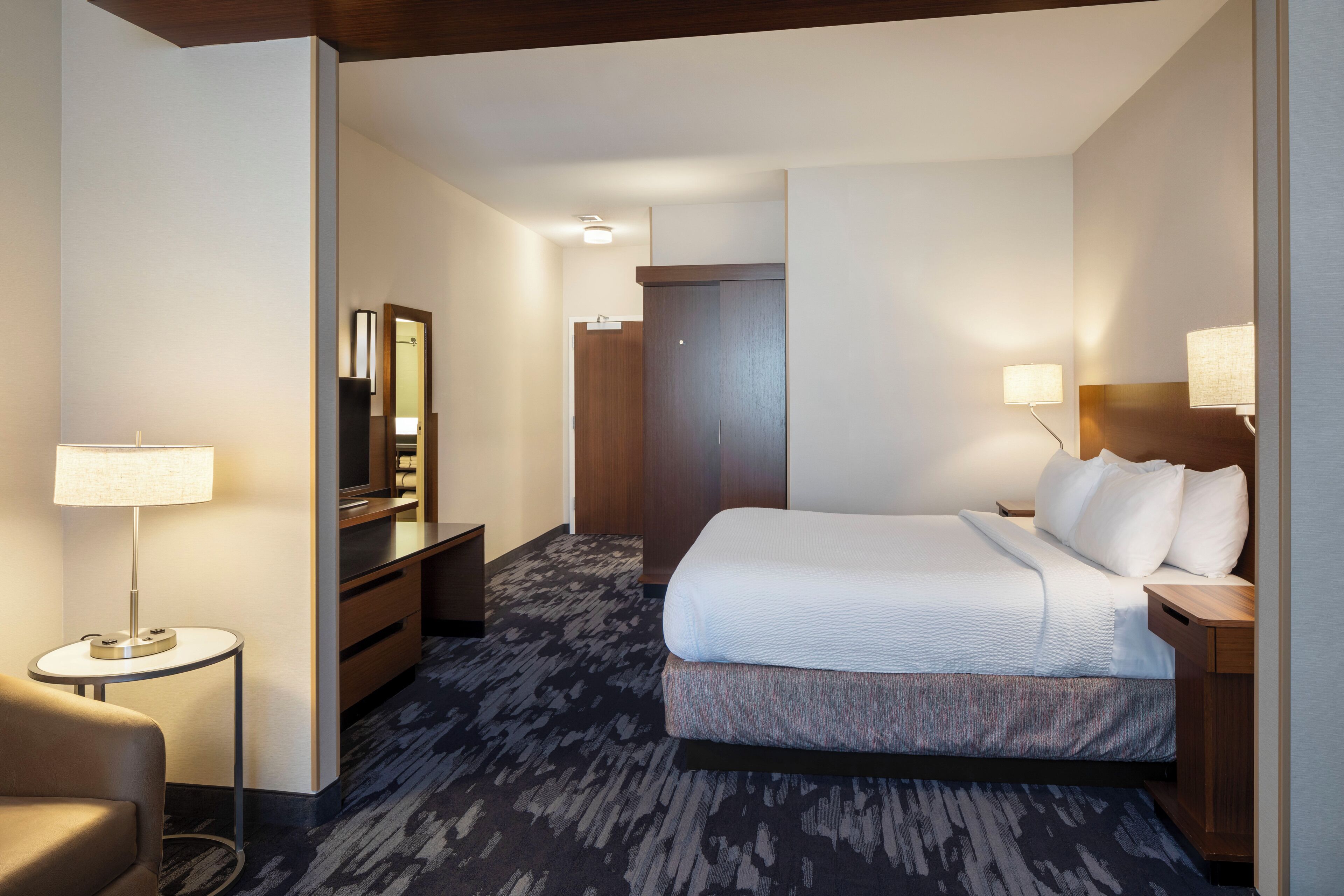 A hotel room with a large bed, a desk, a chair, a mirror, and a lamp.