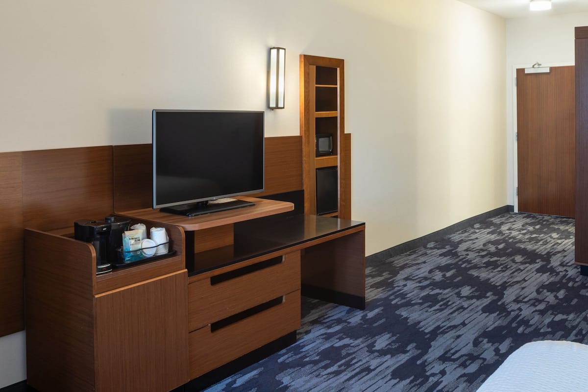 A hotel room with a wooden desk, a flat-screen TV, a small shelf, and a bed.