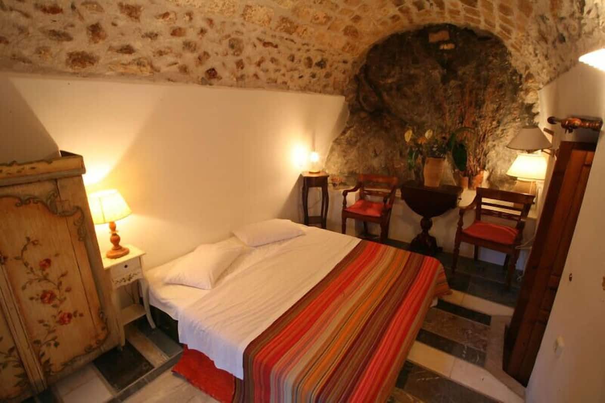 Un dormitorio con pared de piedra, una cama con manta a rayas, dos sillas y dos lámparas de noche.