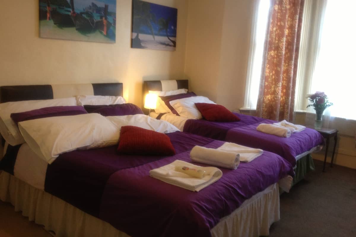 A hotel room with two beds, each with purple bedding and white pillows. There are two paintings on the wall, a window with curtains, and a small table with a vase of flowers.