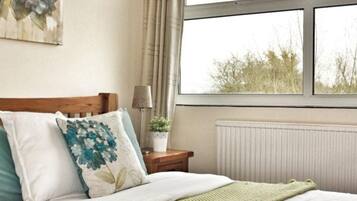 A bedroom with a wooden headboard, a bedside table with a lamp, a potted plant, and a view of trees through a window.