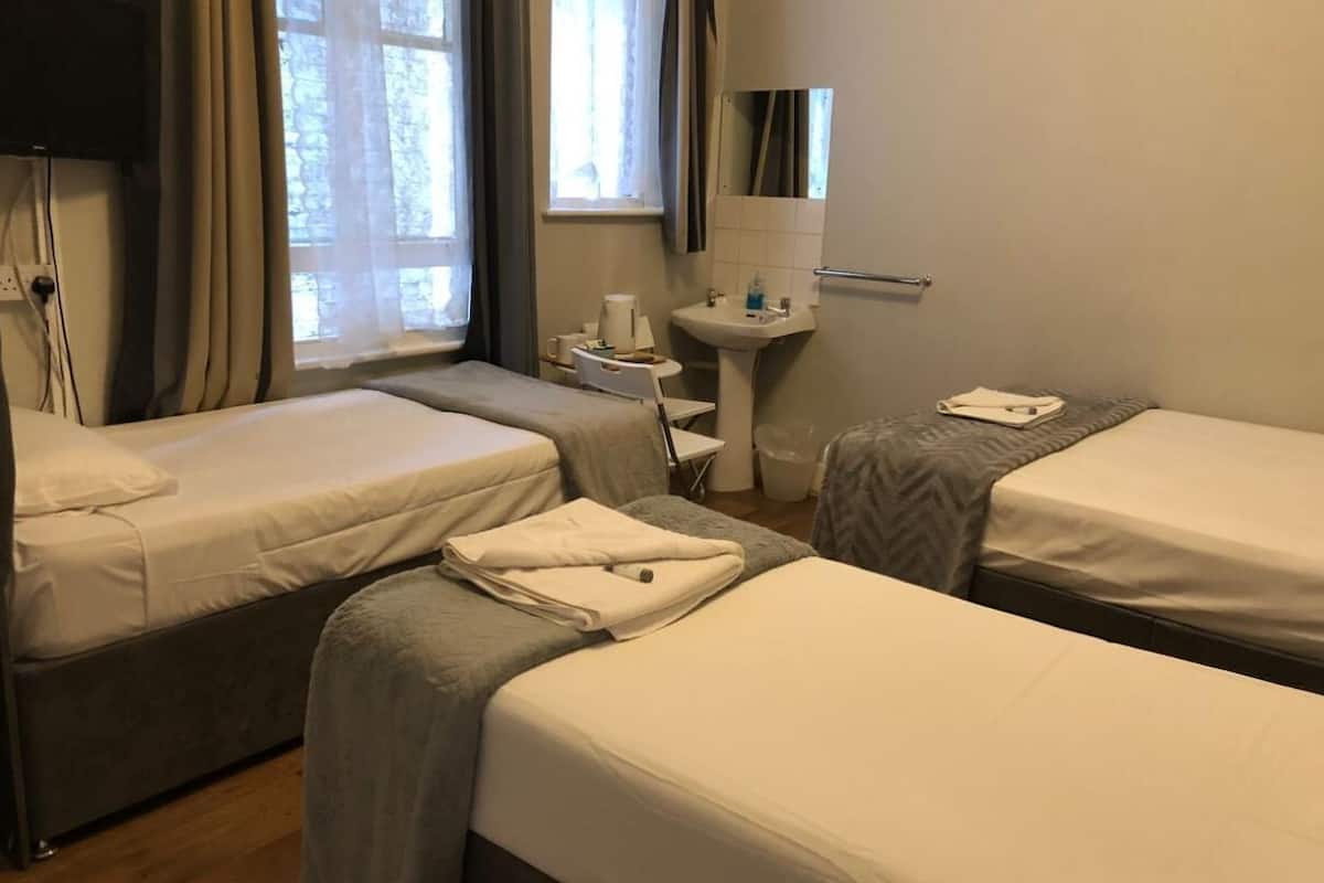 A hotel room with two single beds, a small bedside table with a lamp, a window with curtains, and a wall-mounted mirror.