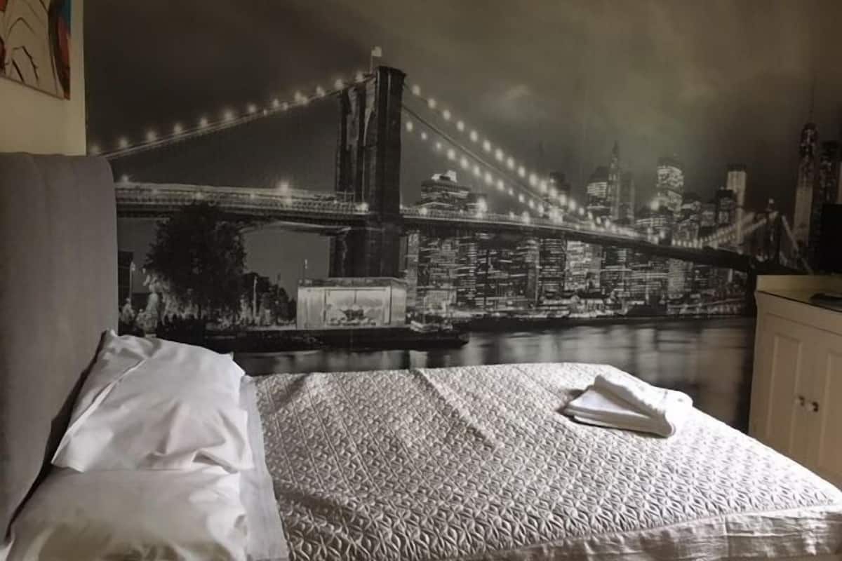 A bedroom with a bed, a white pillow, and a blanket. There is a wall mural of a bridge and cityscape.