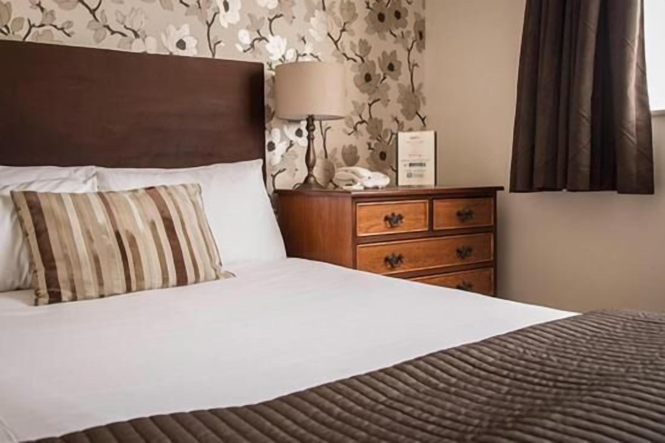 A neatly made bed with a striped pillow, a wooden nightstand with a lamp, and a floral wallpaper.