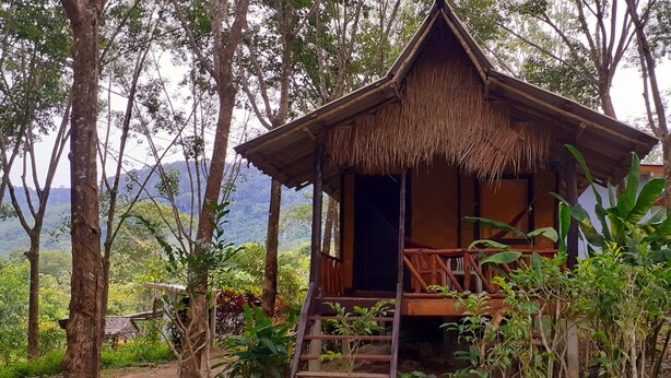 A rustic wooden cabin with a thatched roof, surrounded by lush greenery and trees. - Lanta Maikeaw Bungalow (Ko Lanta)