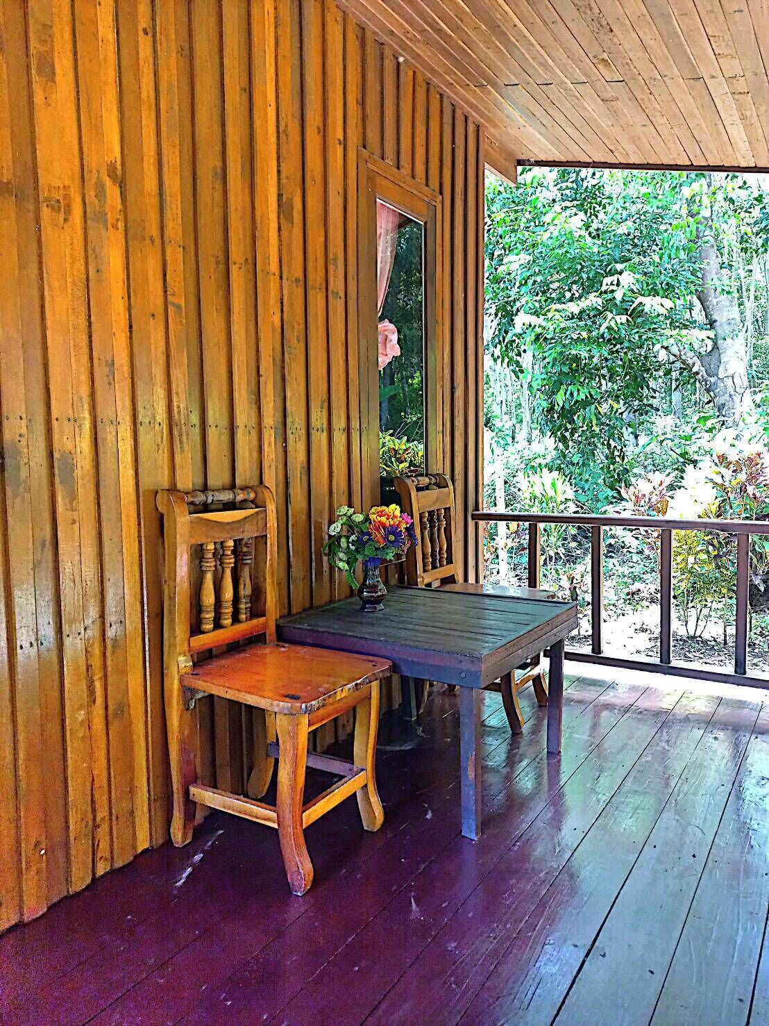 Un porche de madera con una mesa y sillas, un florero con flores y vistas al verdor exterior.