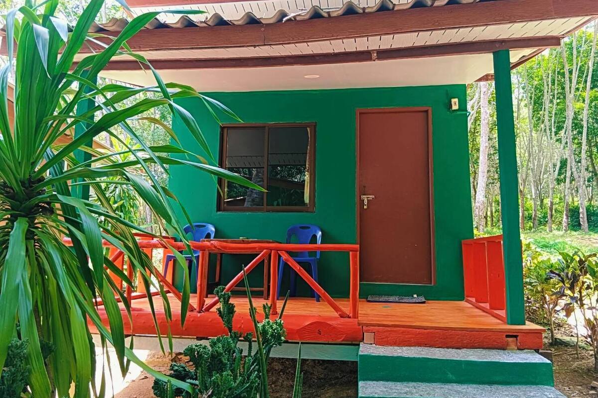 Un bâtiment vert avec un toit rouge, une porte et deux chaises bleues à l’extérieur.