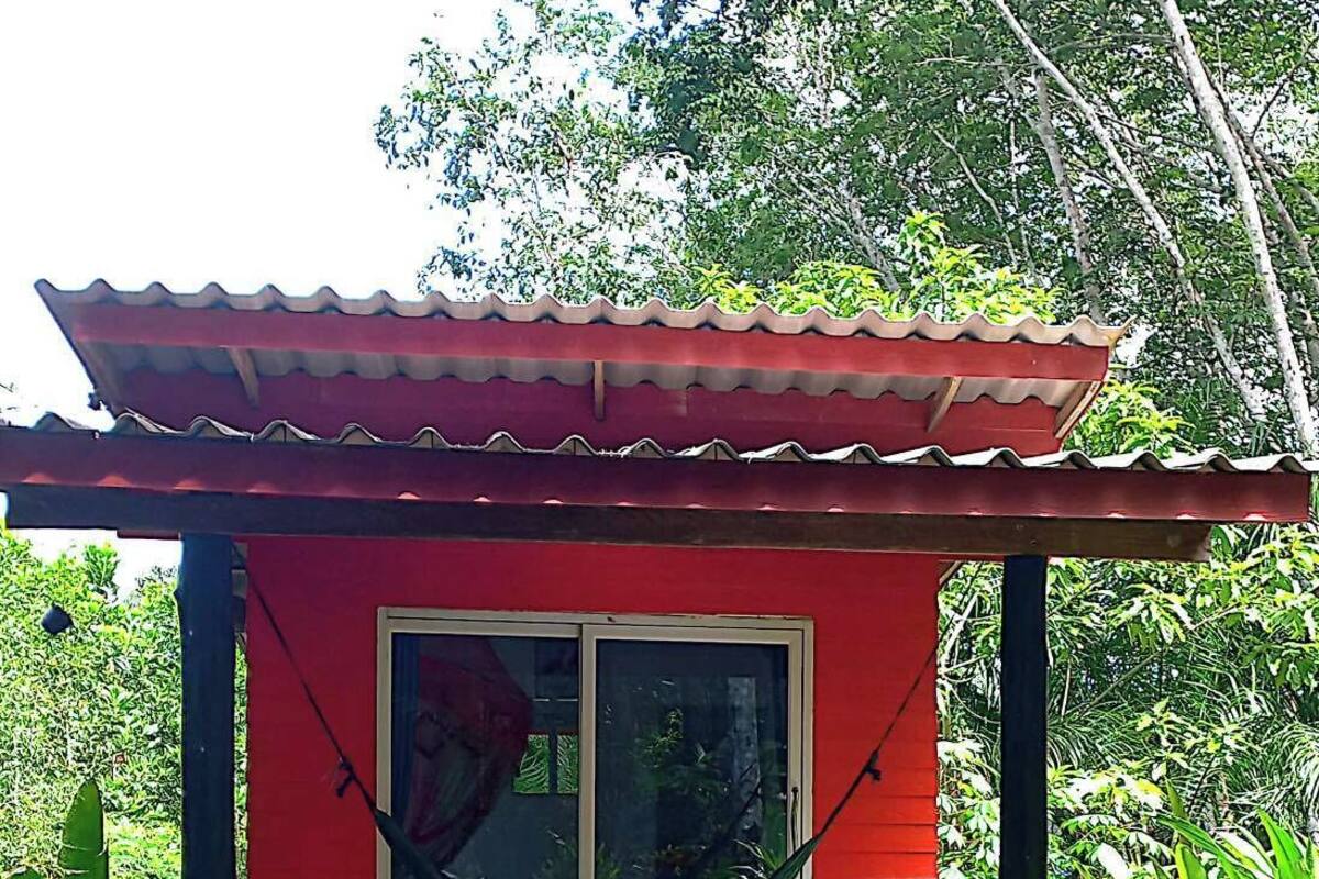 Un bâtiment rouge avec un toit en tuiles et une porte en verre, entouré d’une végétation luxuriante.
