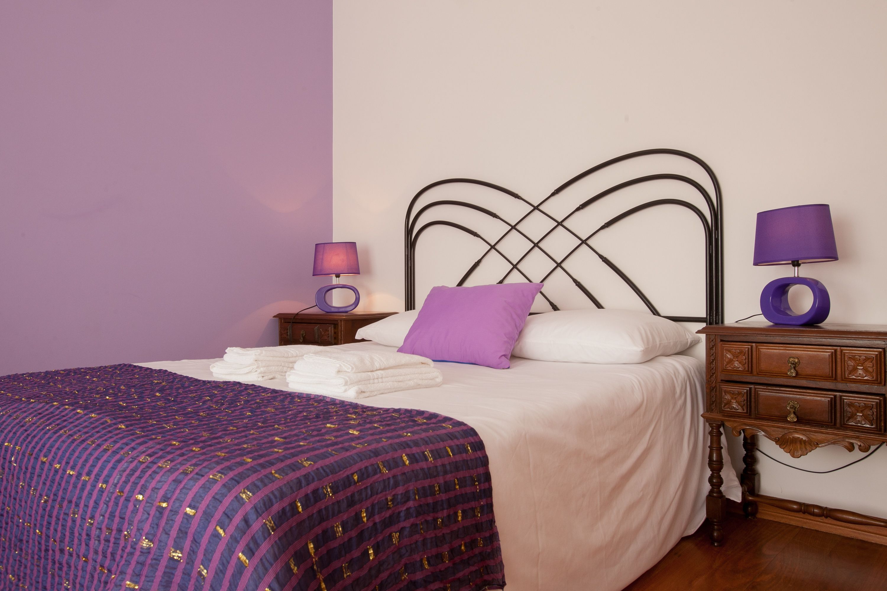 A neatly made bed with a purple and gold patterned bedspread, a white comforter, and a purple pillow. There are two bedside tables with lamps and a wooden nightstand with a lamp.