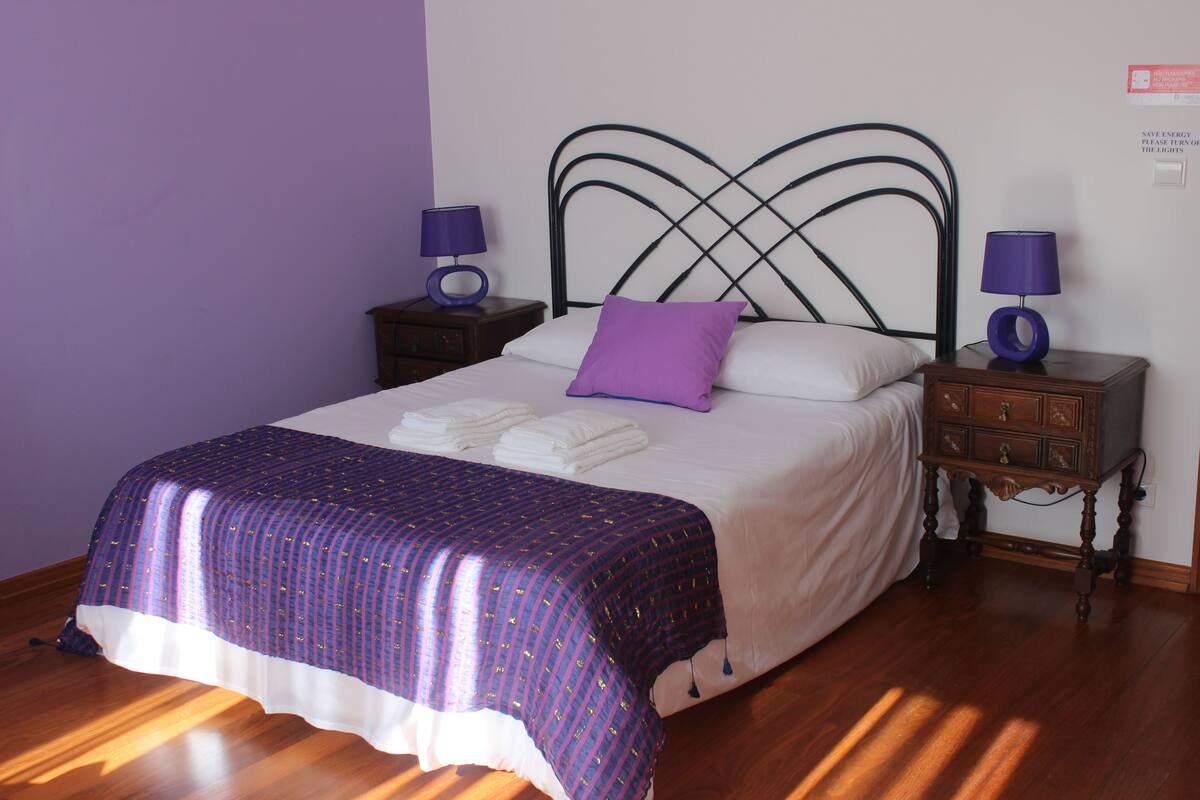 A neatly made bed with a purple and white bedspread, two bedside tables with lamps, and a wall-mounted light switch.