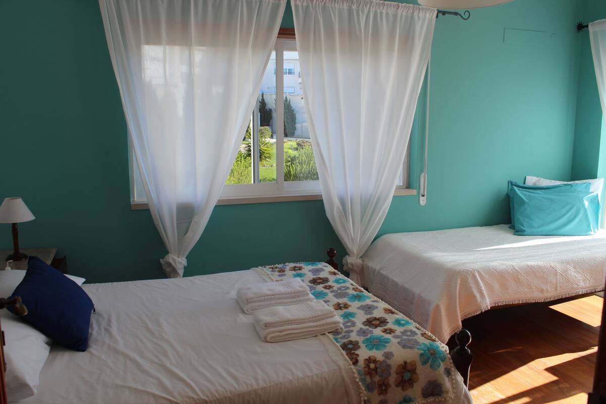 A room with two beds, a window with curtains, and a view of greenery outside.