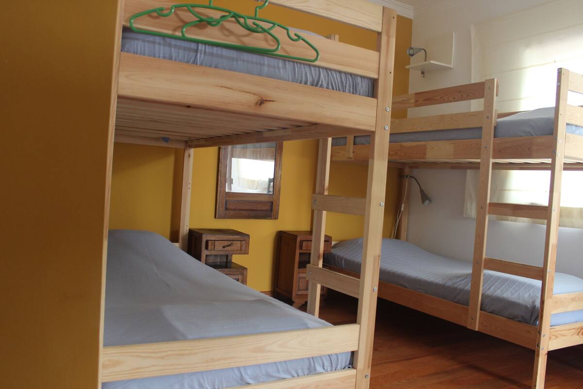 A bunk bed room with wooden frames, a small bedside table, and a window with curtains.