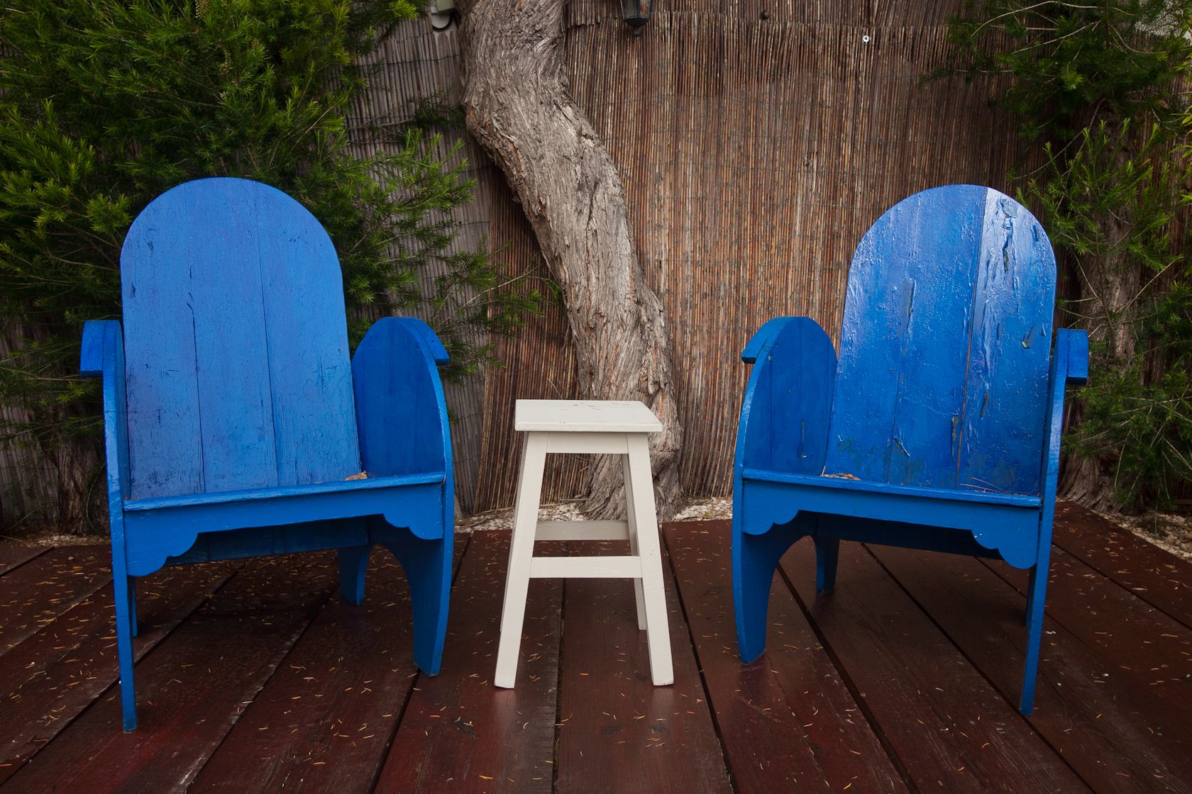 Dos sillas Adirondack azules y un taburete blanco pequeño sobre una terraza de madera, con un árbol al fondo.