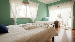 Une chambre avec deux lits, du linge de lit blanc et une vue sur l’extérieur à travers des rideaux transparents.