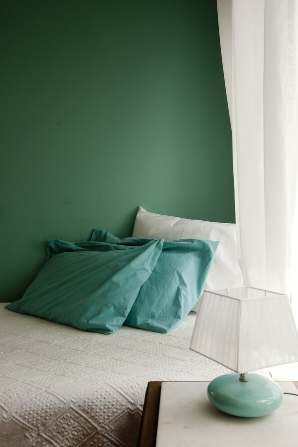 Un lit avec une literie aux couleurs turquoise et blanche, une lampe blanche posée sur une table de chevet et un mur vert.