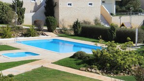 Une piscine dotée d’une terrasse carrelée, entourée de pelouse et d’arbustes bien entretenus, et jouxtant un bâtiment à la façade en pierre et doté de balcons.