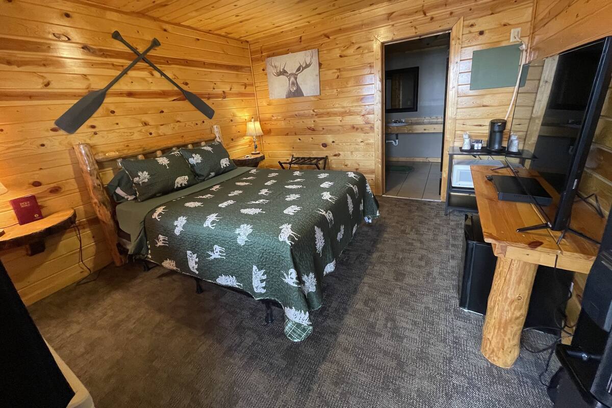 A wooden cabin room with a bed, a desk, a chair, a lamp, and a painting on the wall.