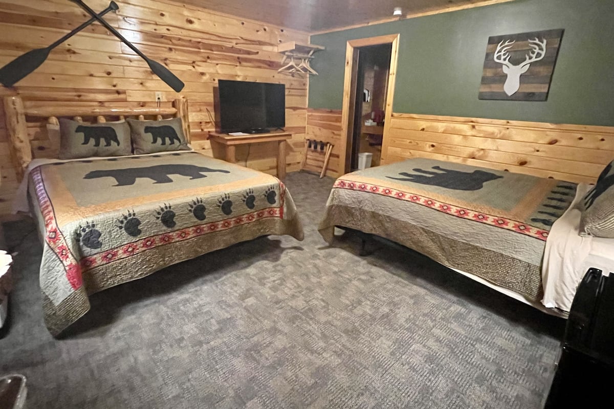 A cabin-style room with two beds, each with a bear motif, a television on a wooden table, and a deer wall art.