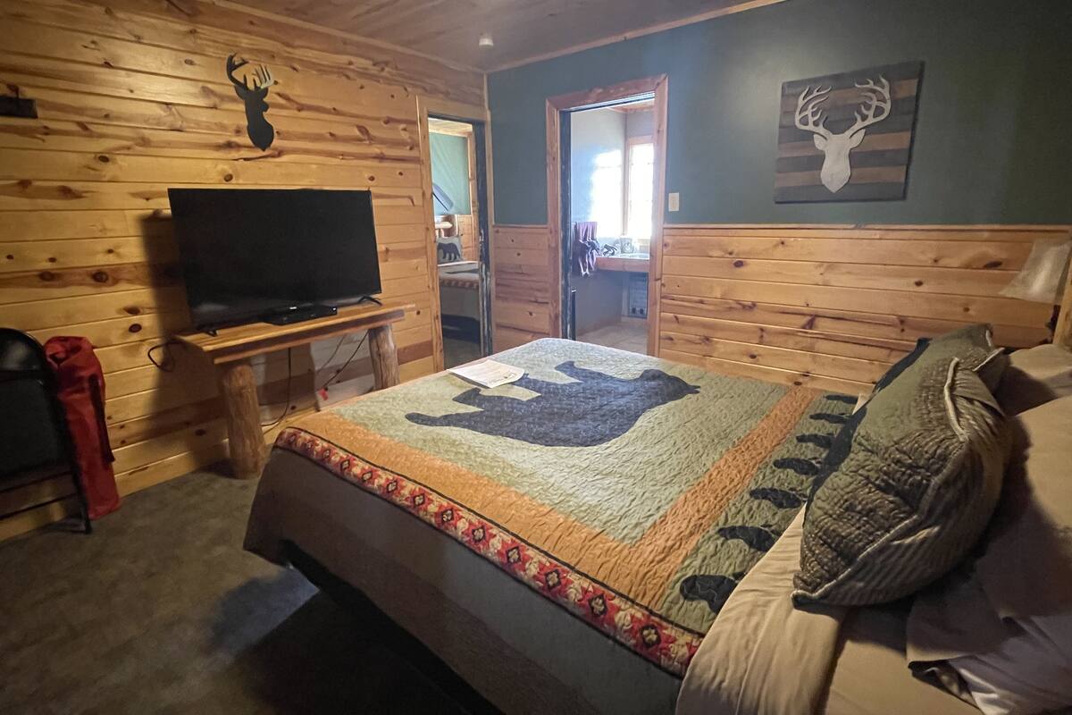 A cozy room with a wooden ceiling and walls, a bed with a patterned quilt, a television on a wooden stand, and a door leading to another room.