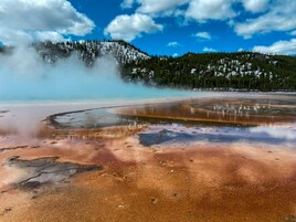 Un área geotérmica con depósitos minerales y vapor ascendiendo del suelo, rodeada de montañas boscosas.