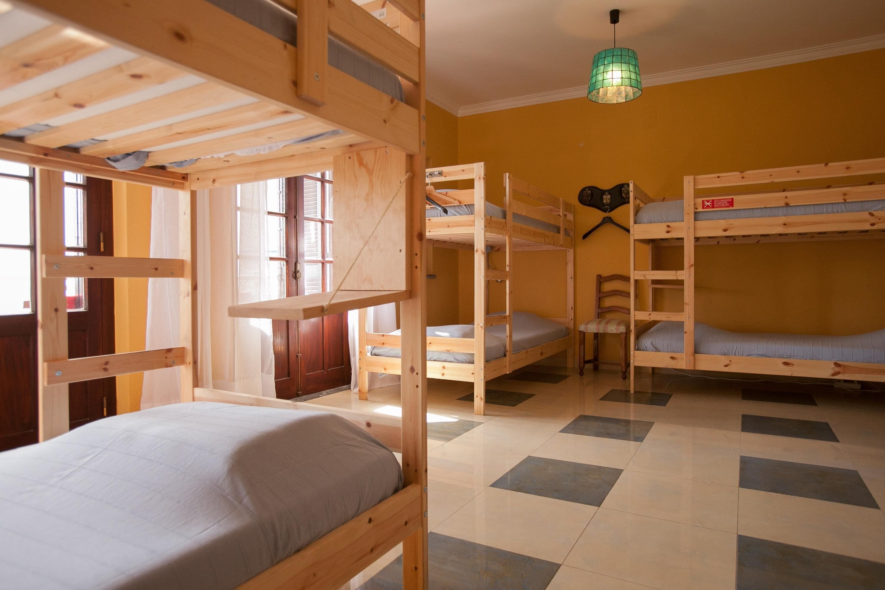 A room with bunk beds, a window with curtains, a green hanging light, and a tiled floor.