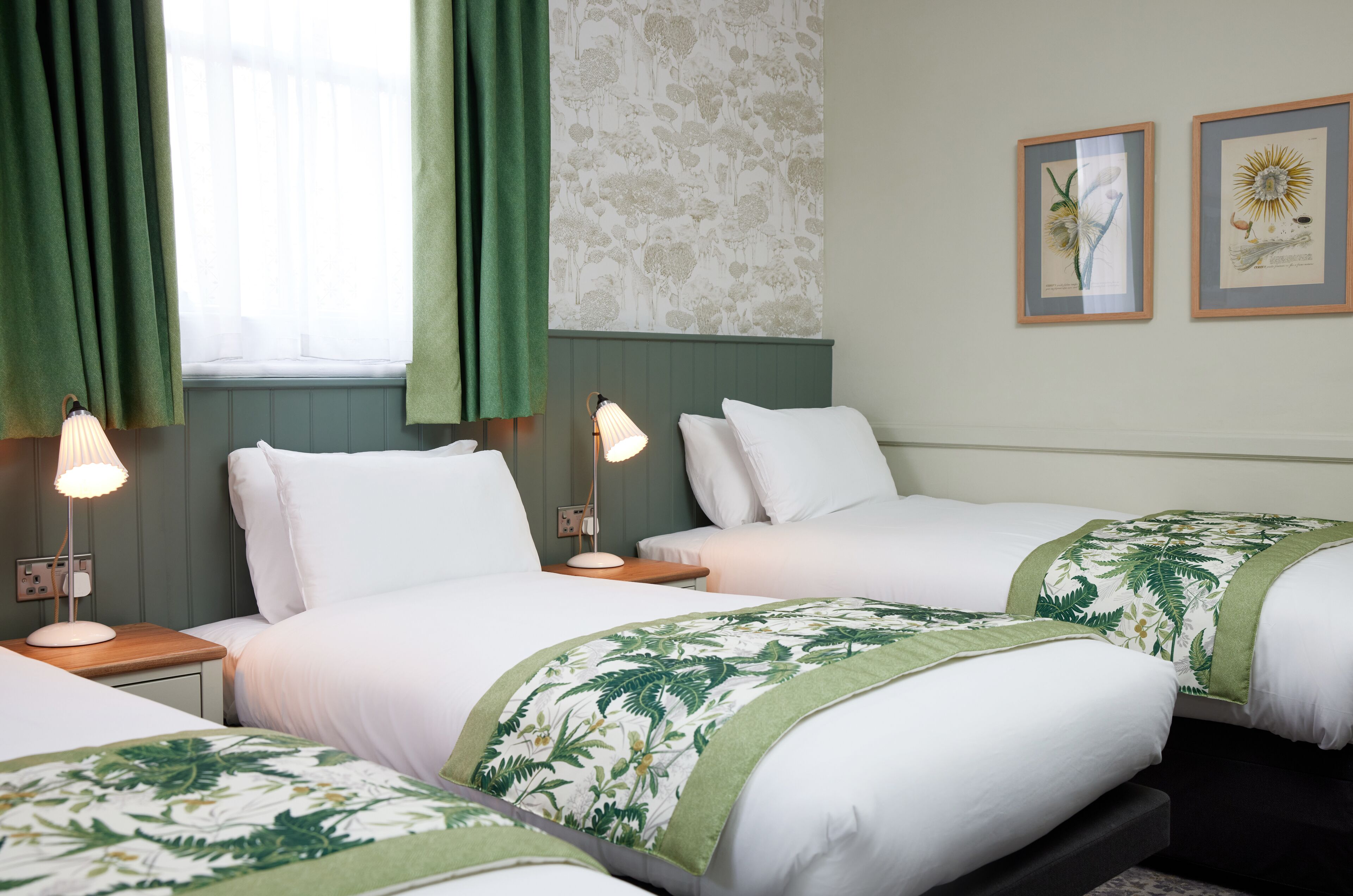 Habitació d'hotel amb dos llits individuals, un edredó floral verd i blanc, una petita taula de nit amb una làmpada i una finestra amb cortines verdes.