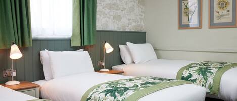 Habitació d'hotel amb dos llits individuals, un edredó floral verd i blanc, una petita taula de nit amb una làmpada i una finestra amb cortines verdes.