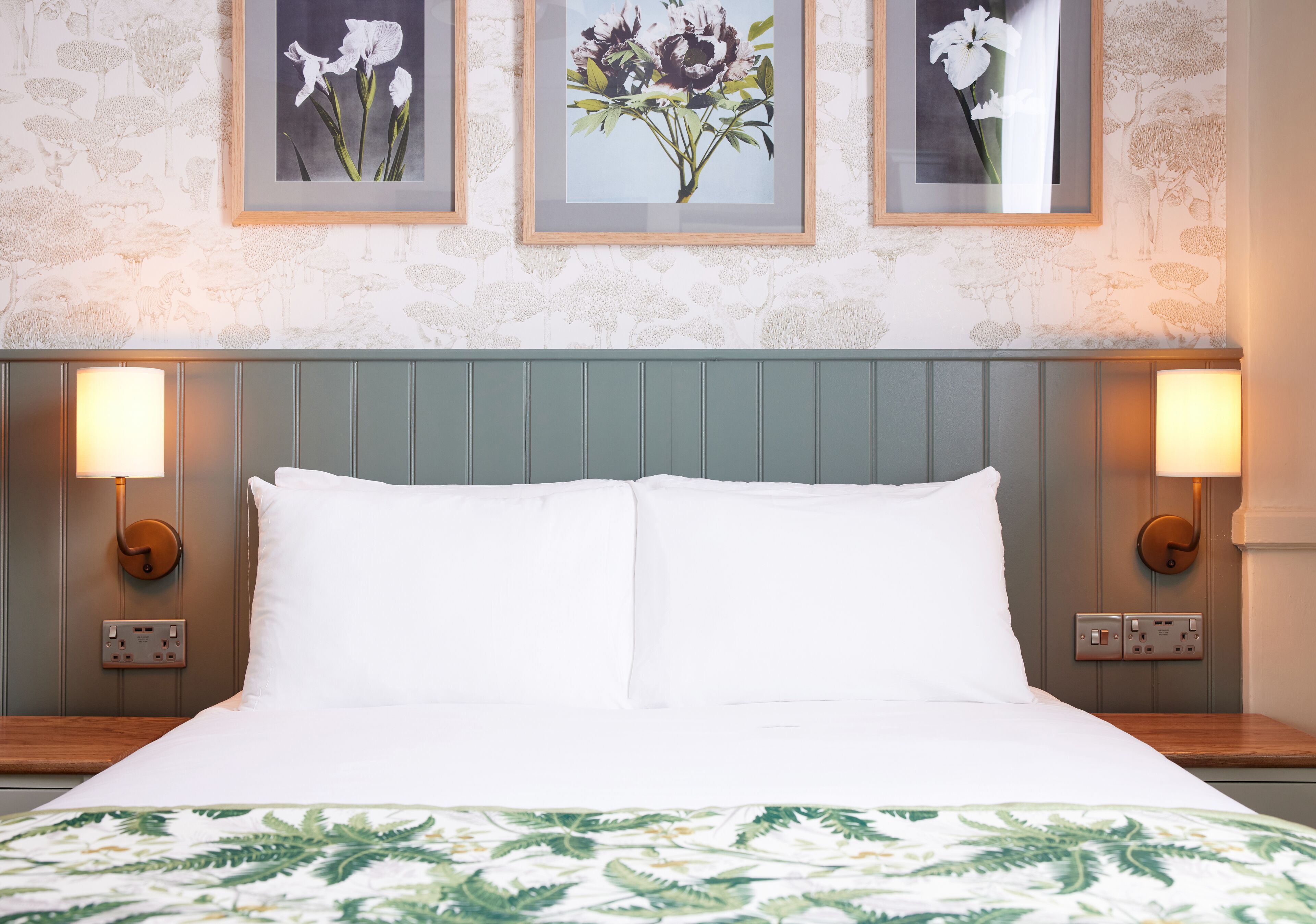 Een hotelkamer met een bed, twee wandlampen en drie florale foto's in lijstjes aan de muur.