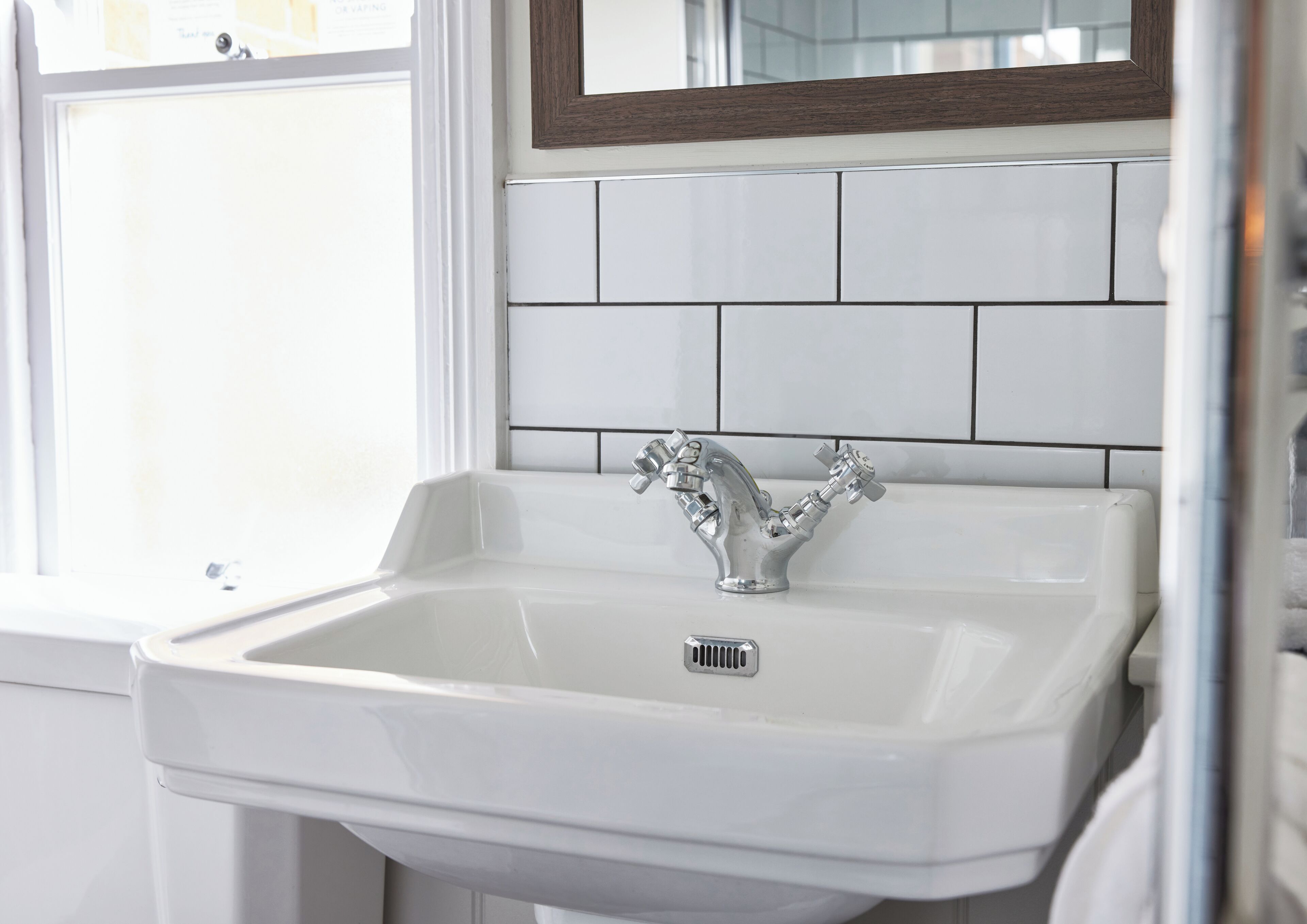 Un lavabo blanc dans une salle de bain, doté d’un robinet moderne, des murs recouverts de carreaux blancs et d’une fenêtre avec des stores.