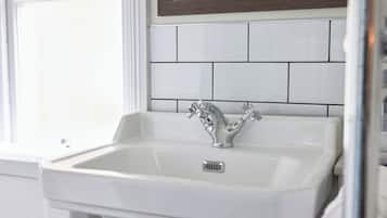 Un lavabo blanc dans une salle de bain, doté d’un robinet moderne, des murs recouverts de carreaux blancs et d’une fenêtre avec des stores.