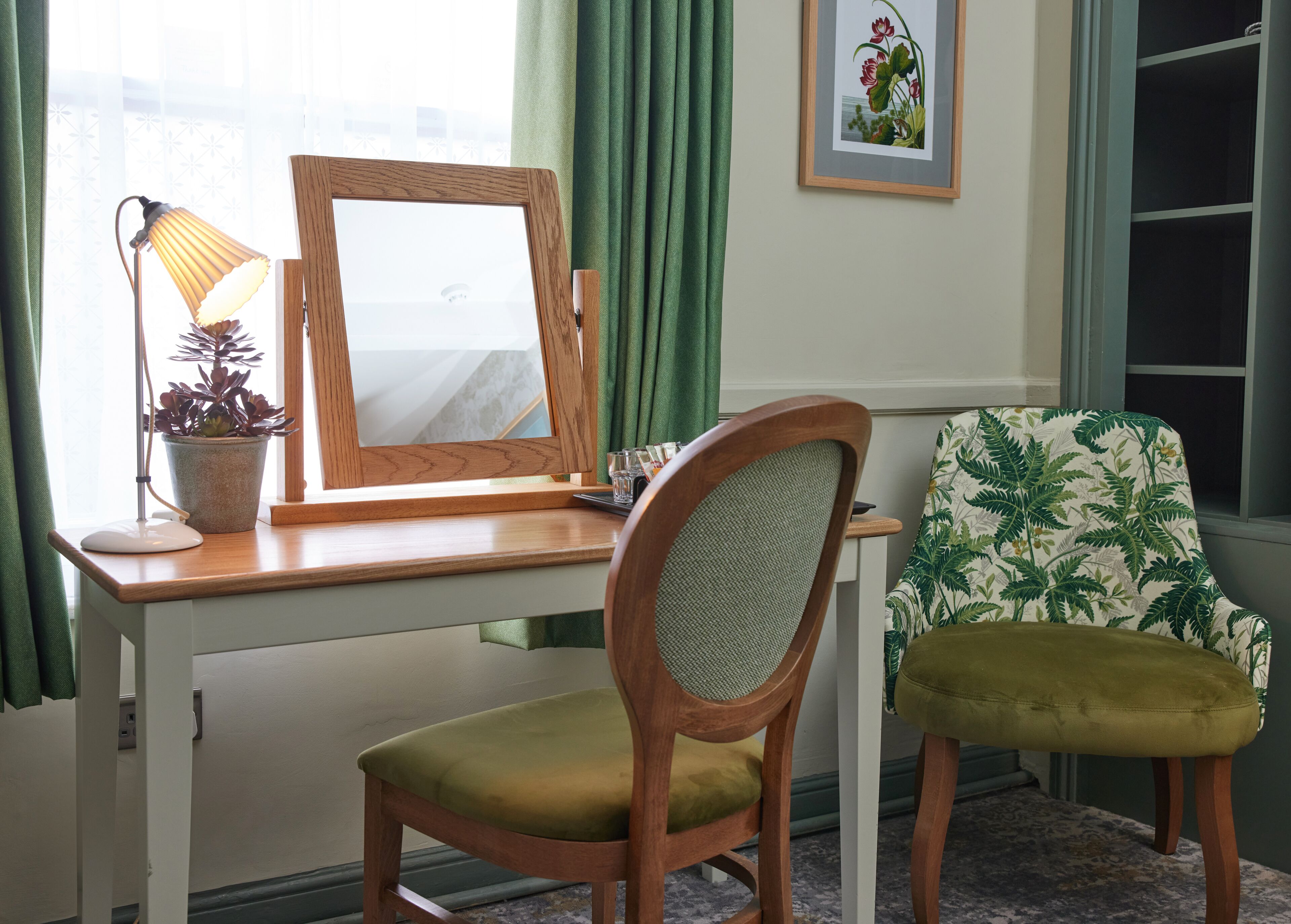 Une pièce avec un bureau en bois, une chaise, une plante en pot, un tableau encadré et une fenêtre avec des rideaux.
