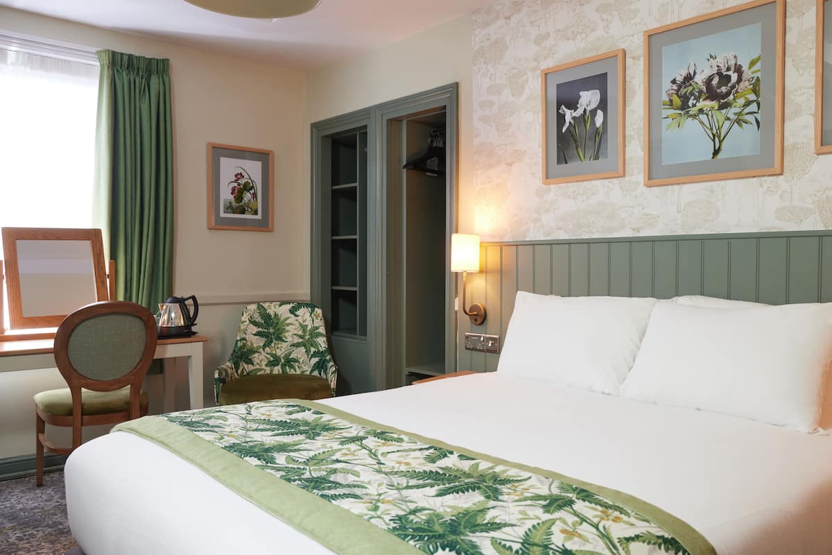 A hotel room with a bed, a desk, a chair, a wardrobe, and framed floral pictures on the wall.