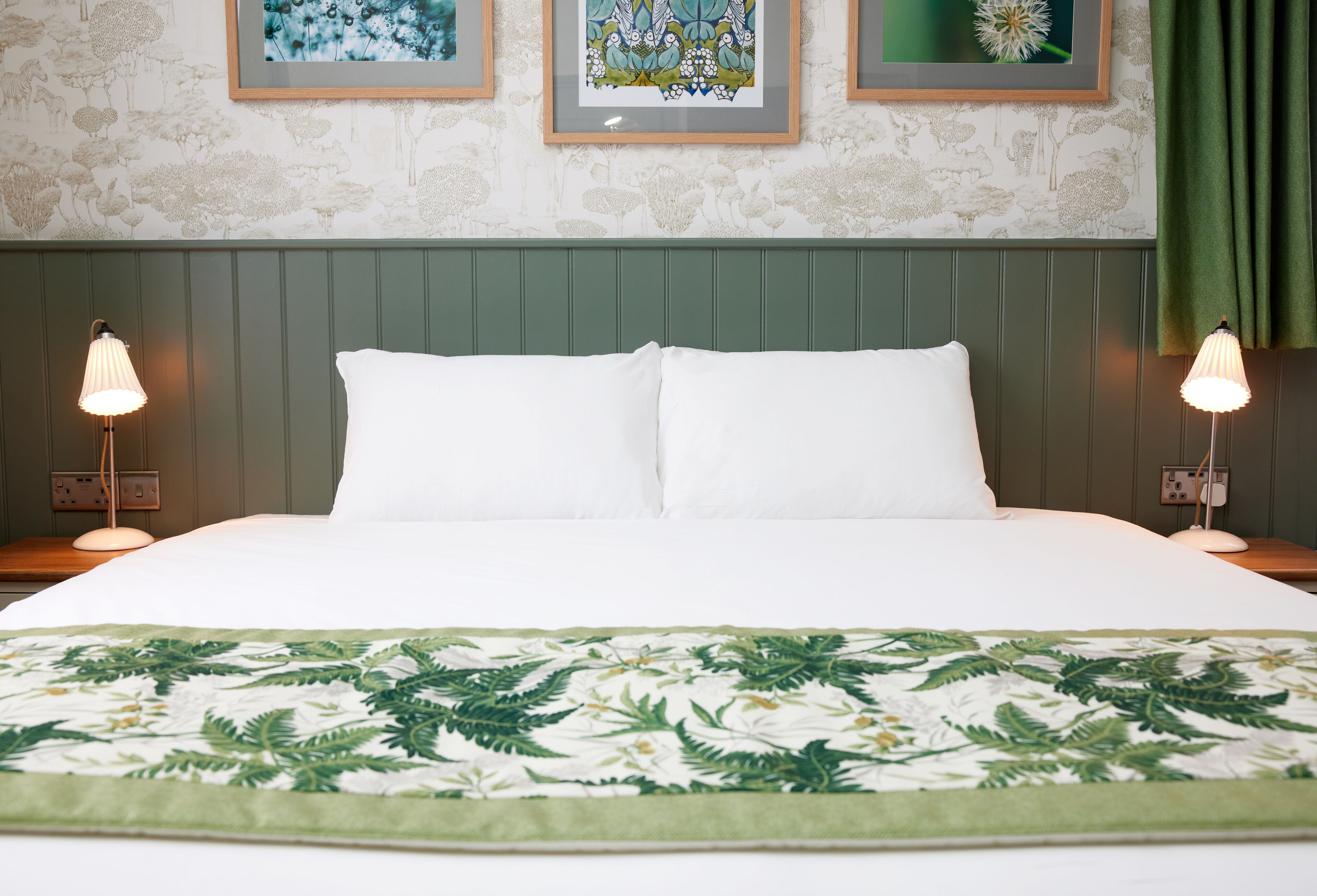 Una cama bien hecha con sábanas blancas, una colcha con estampado de hojas verdes y dos lámparas de noche.