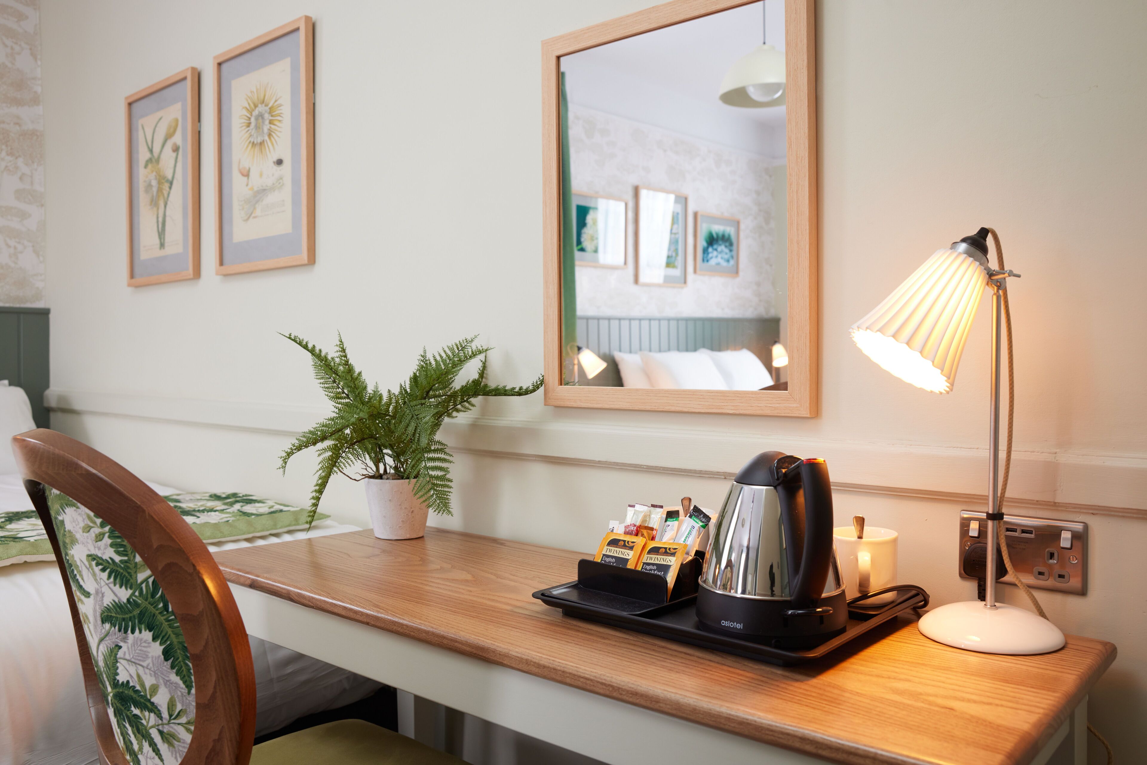 A hotel room with a wooden desk, a potted plant, a mirror, framed artwork on the wall, and a bedside lamp.