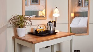 A hotel room with a wooden table, a potted plant, a tray with coffee cups and a kettle, and a lamp.