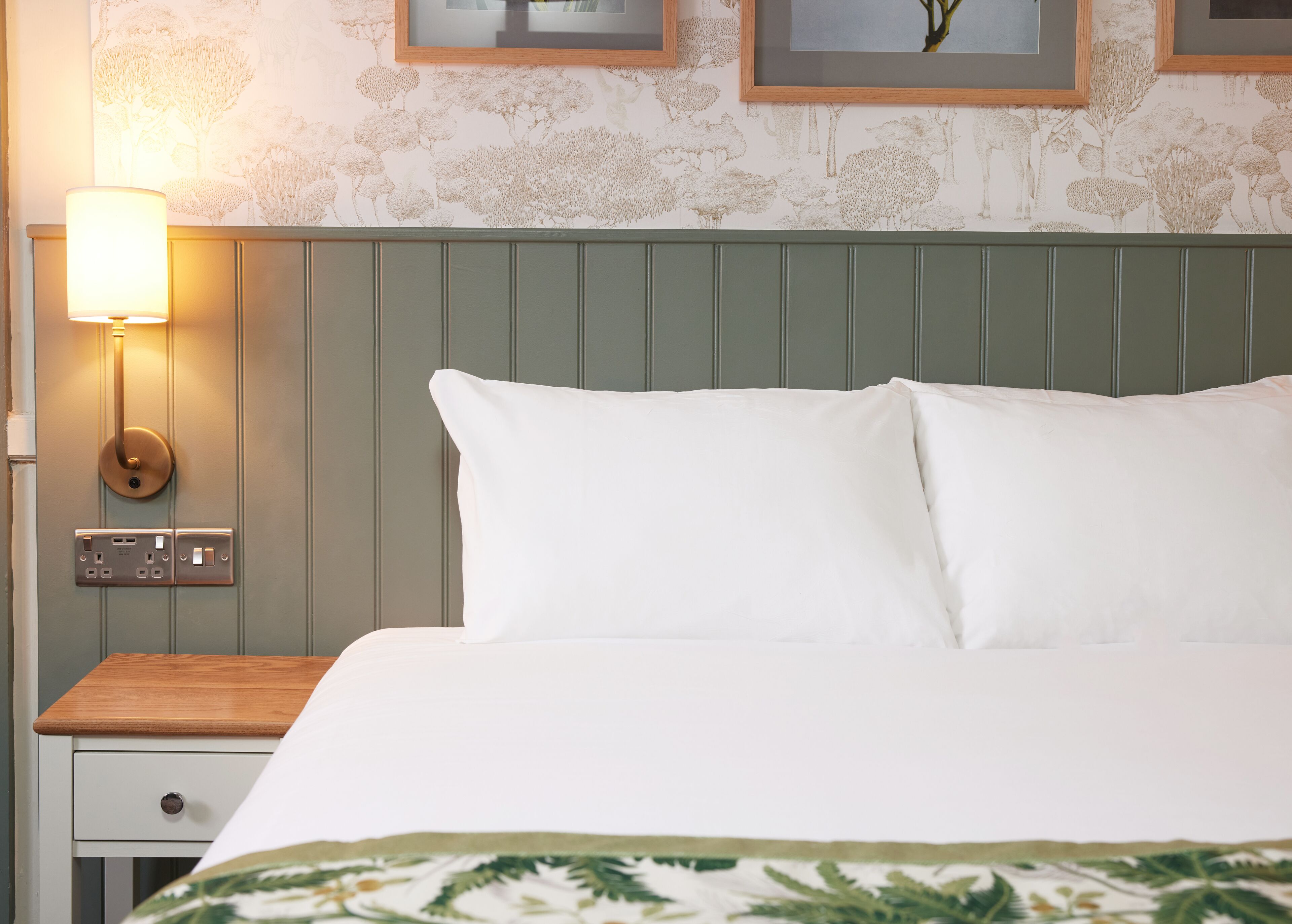 Una cama bien hecha con ropa blanca y un cabecero verde, una mesita de noche con una lámpara, y una pared con impresiones botánicas enmarcadas.