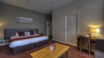A hotel room with a bed, a desk, a chair, a table, and a wall-mounted lamp.