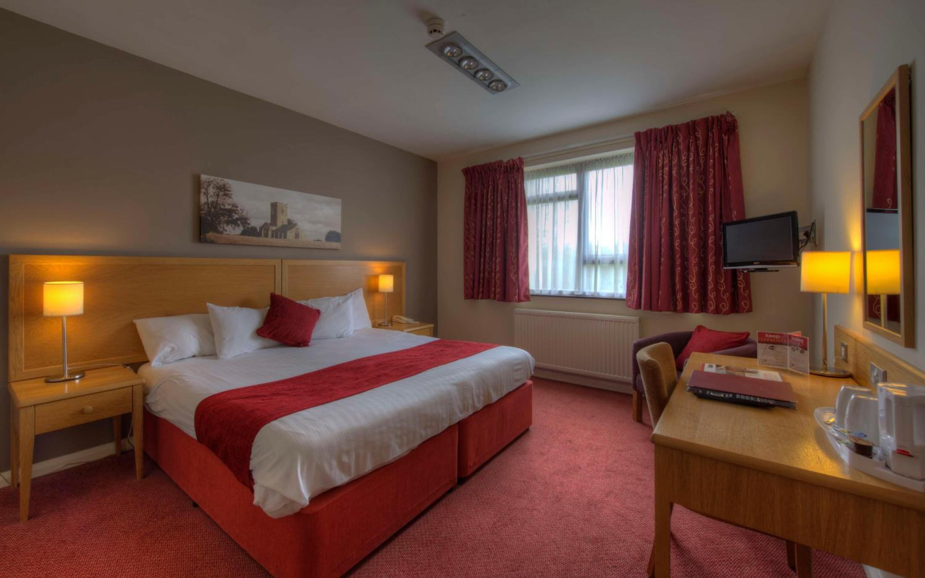 A hotel room with a large bed, two bedside lamps, a desk with a chair, a television, and a window with curtains.